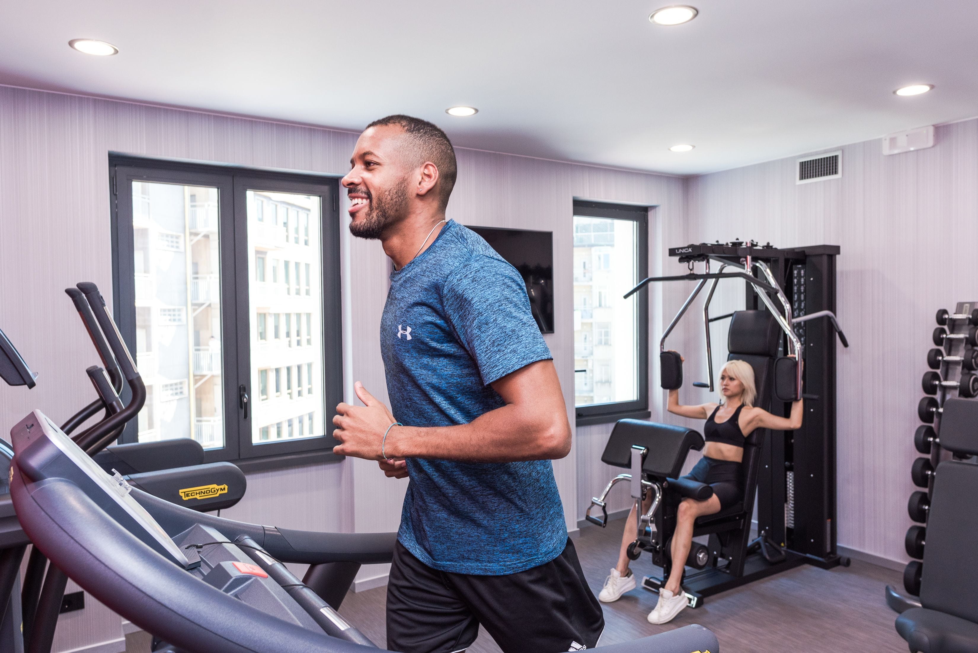 Un gimnasio con cintas de correr, una mujer ejercitándose en una máquina y un hombre corriendo en una cinta.