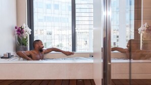 Ein Mann entspannt in einer Badewanne mit Blick auf eine Stadtlandschaft durch große Fenster.