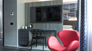Quarto de hotel moderno com televisão de tela plana na parede, escrivaninha com chaleira, cadeira preta e cadeira vermelha.