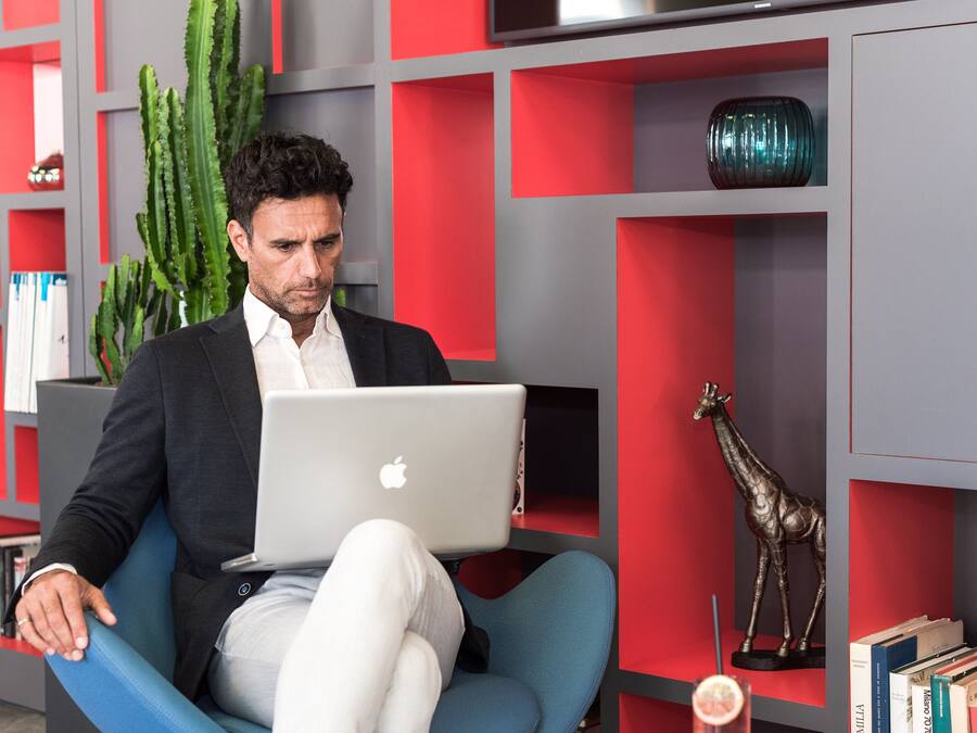Une personne est assise sur une chaise bleue, un ordinateur portable sur les genoux, entourée d’étagères avec des panneaux rouges et gris, d’un cactus et d’une statue de girafe.