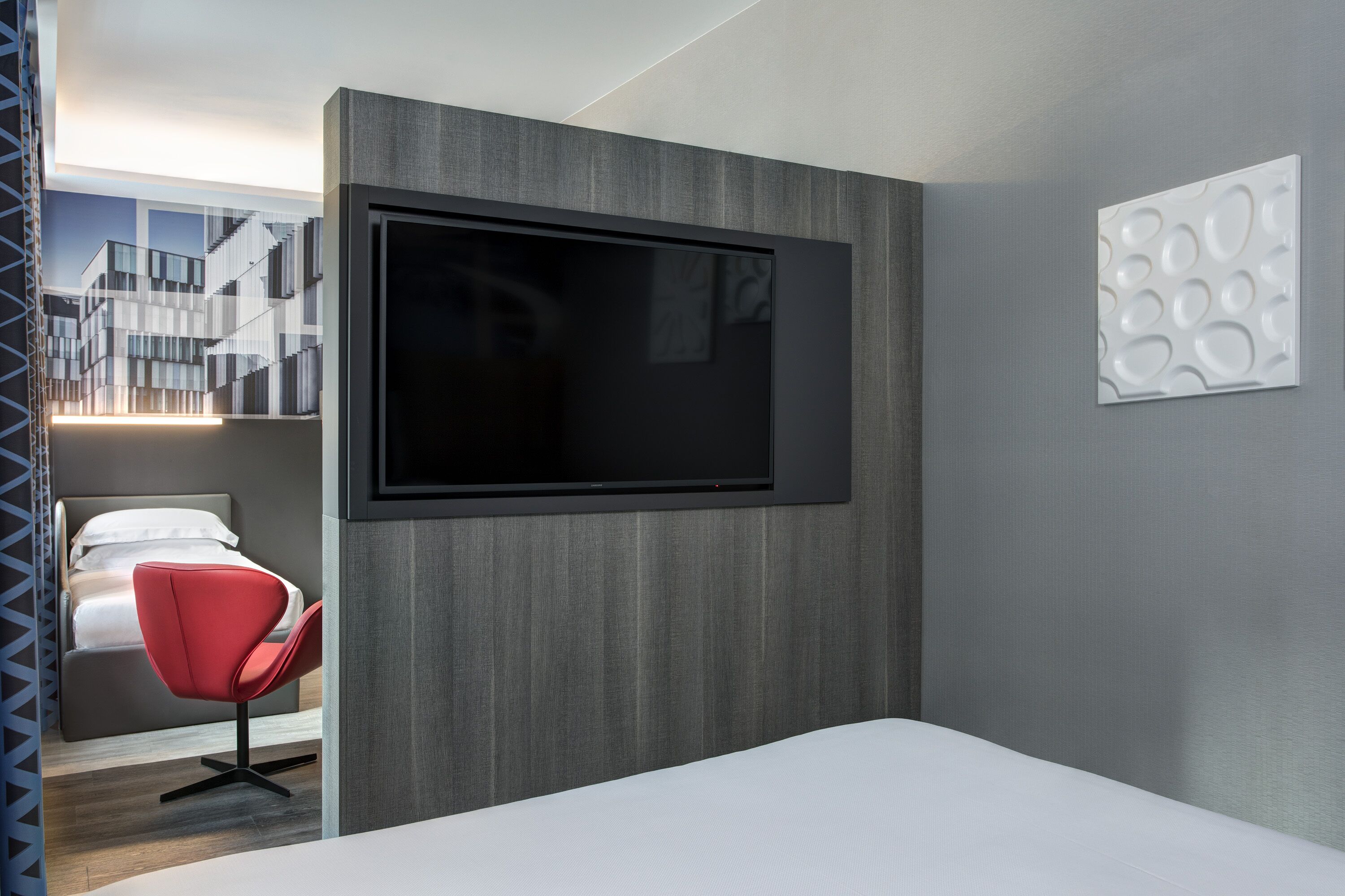 Une chambre d’hôtel moderne avec un grand téléviseur à écran plat fixé au mur, un fauteuil rouge et un lit.