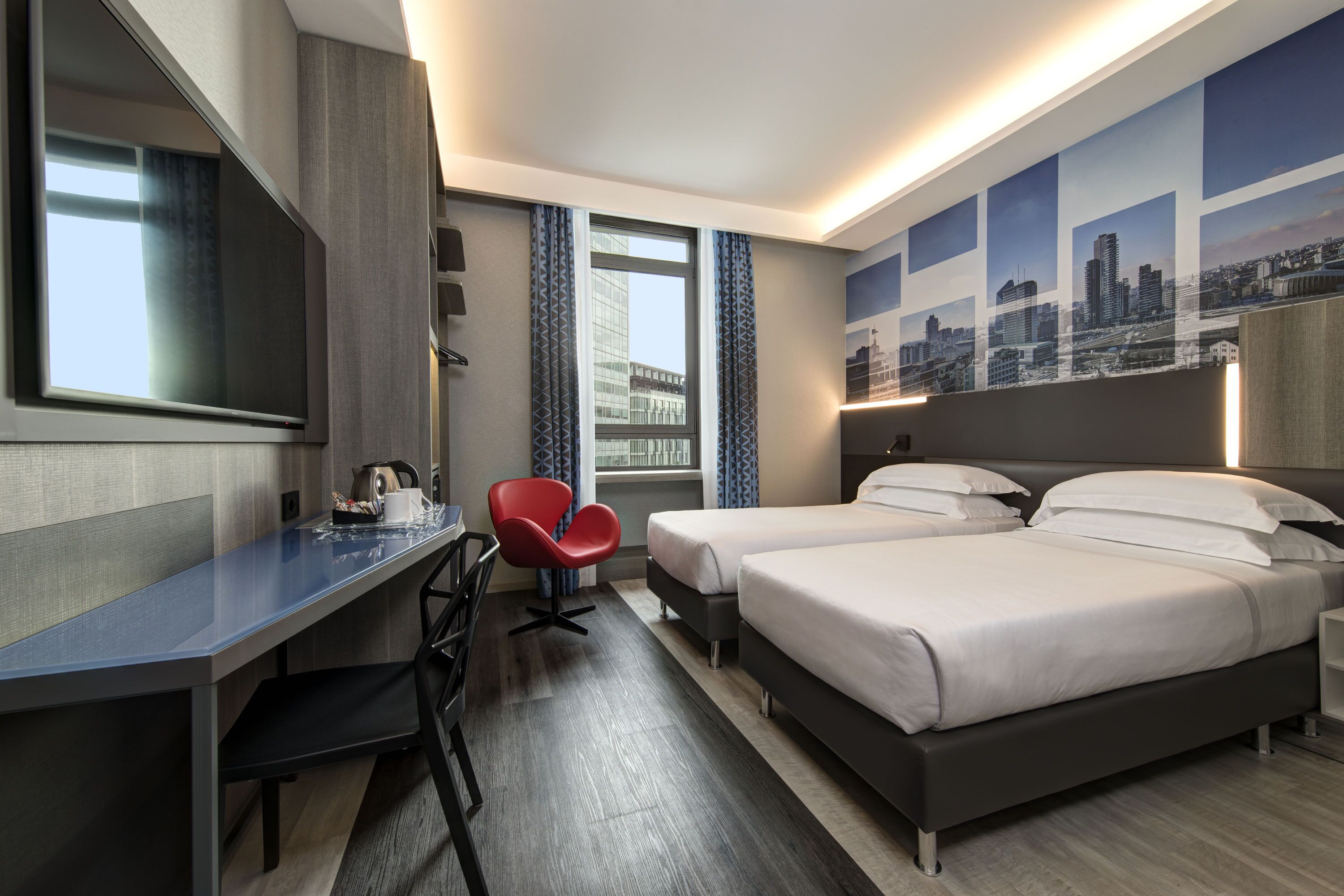 Habitación de hotel con escritorio, dos camas, una silla roja, un ventanal grande y una obra de arte con motivos urbanos en la pared.