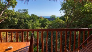 A wooden deck with a railing, a table, and two chairs. The deck overlooks a lush green landscape with trees and a view of the ocean.