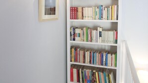 Ein Flur mit einem weißen Bücherregal voller Bücher, einem gerahmten Bild an der Wand und einer Treppe unter einer Decke mit Holzbalken.