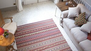 Ein gemütliches Zimmer mit einem weißen Sofa, einem gemusterten Teppich, einem Holznachttisch mit Lampe und einem Fenster mit weißen Vorhängen.
