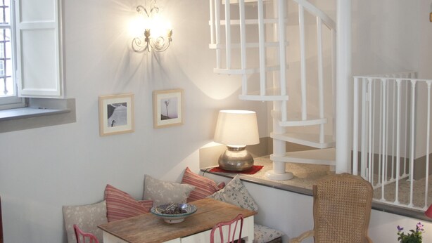 Ein gemütliches Zimmer mit Esstisch, Stühlen, einer Lampe, einem Korbstuhl, einem kleinen Tisch mit Pflanze und einer weißen Wendeltreppe.