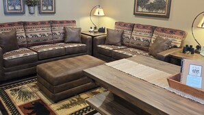 Un salon chaleureux avec un canapé d’angle, un pouf et une table basse en bois. On y trouve des lampes fixées au mur, des œuvres d’art encadrées et une petite table avec une lampe.