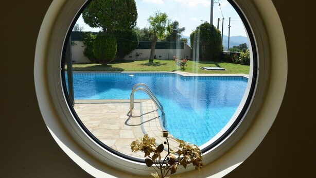 A round window with a view of a swimming pool, a metal ladder, and a small plant. - Villa Xenia (Saronikos)