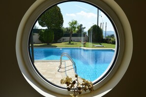 A round window with a view of a swimming pool, a metal ladder, and a small plant. - Villa Xenia (Saronikos)