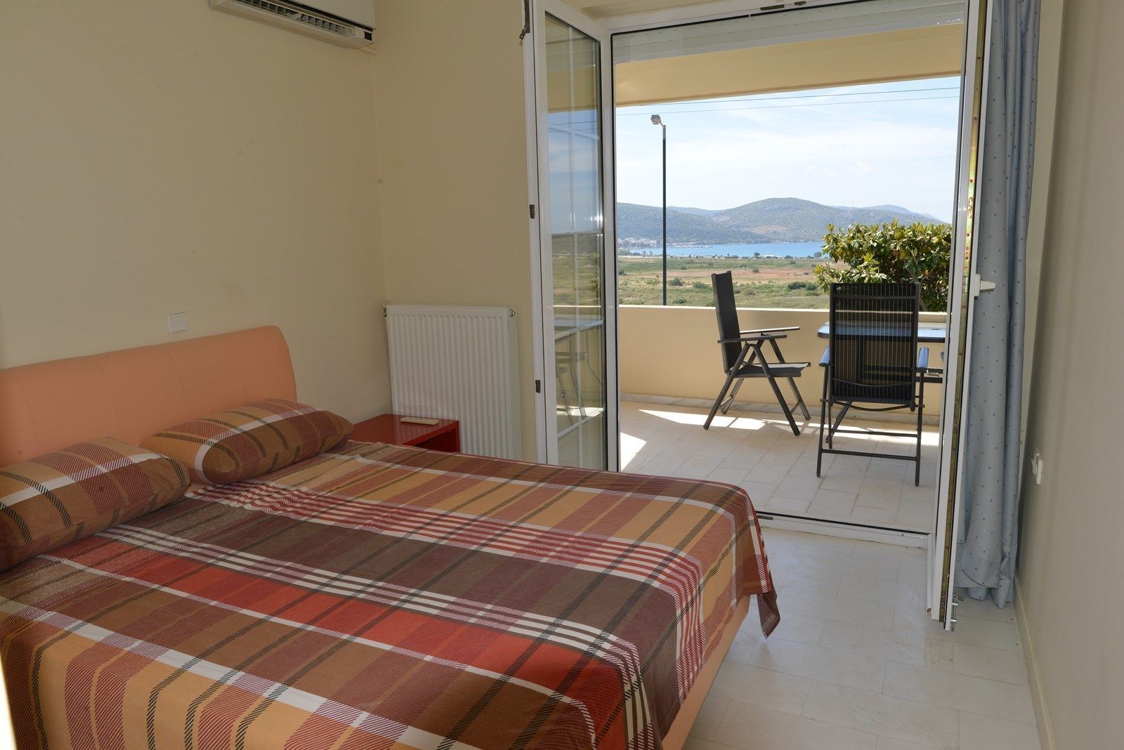 Une chambre à coucher comprenant un lit, une table de chevet, un radiateur et une porte-fenêtre coulissante donnant sur un espace extérieur avec une chaise et une table.