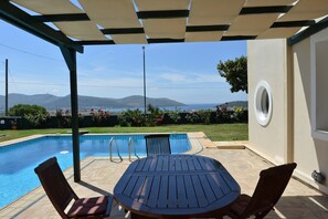 A covered outdoor area with a pool, a table, and chairs, overlooking a scenic landscape with mountains and a body of water. - Villa Xenia (Saronikos)