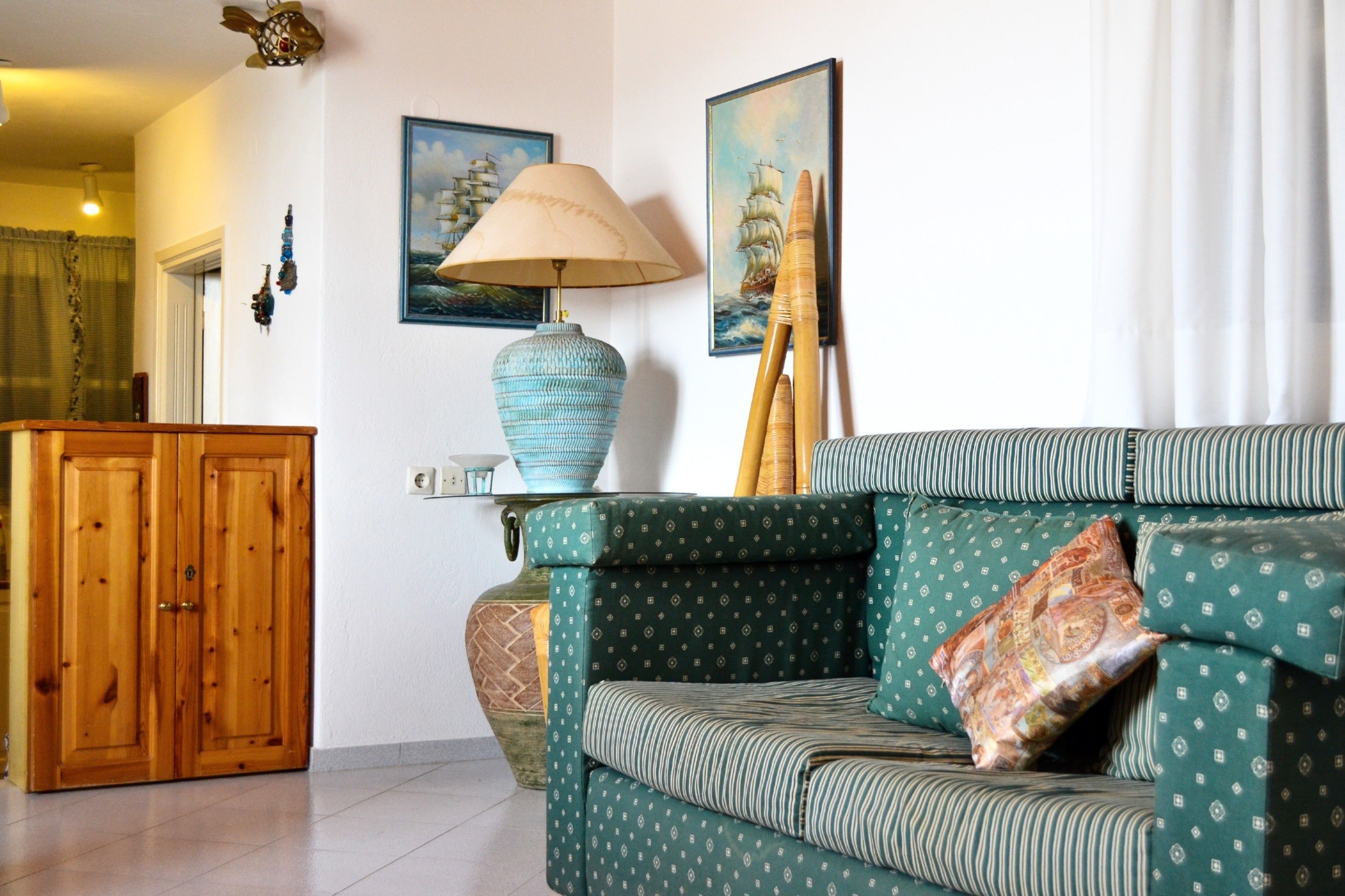 A living room with a green patterned sofa, a wooden cabinet, a blue vase lamp, and two framed paintings on the wall.
