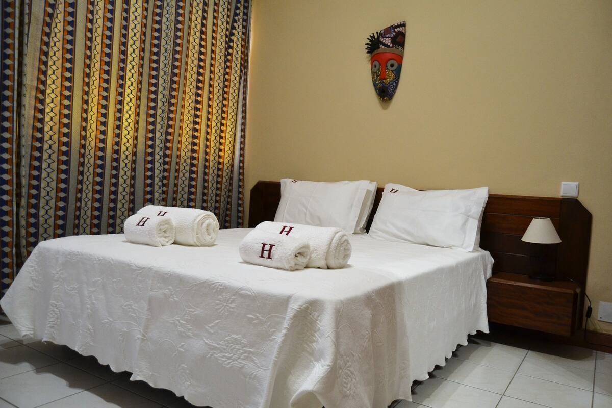 Ein Hotelzimmer mit einem ordentlich bezogenem Bett, zwei Handtüchern mit den Initialen 'H' und einer dekorativen Maske an der Wand, sowie einem Vorhang mit Muster.