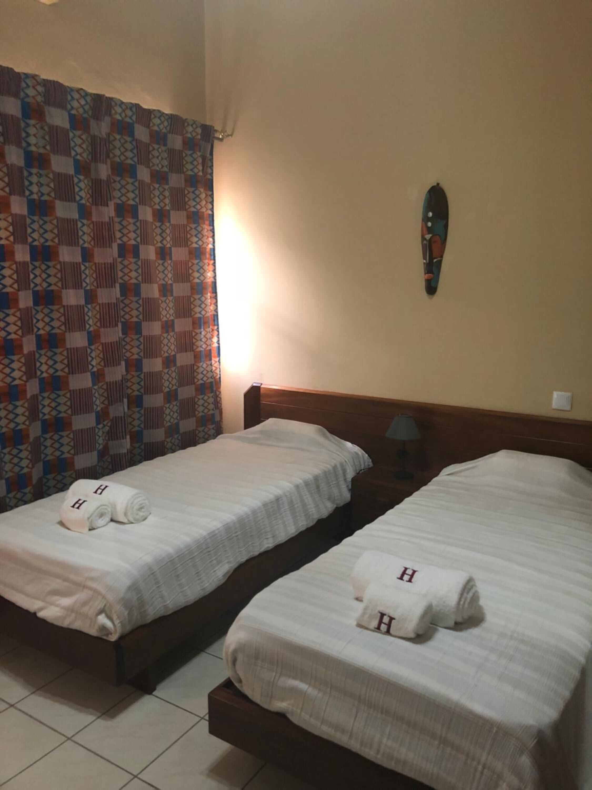 Camera d'albergo con due letti, ognuno con una coperta bianca e asciugamani con la lettera 'H'. Una tenda decorata a un lato e una decorazione a parete.