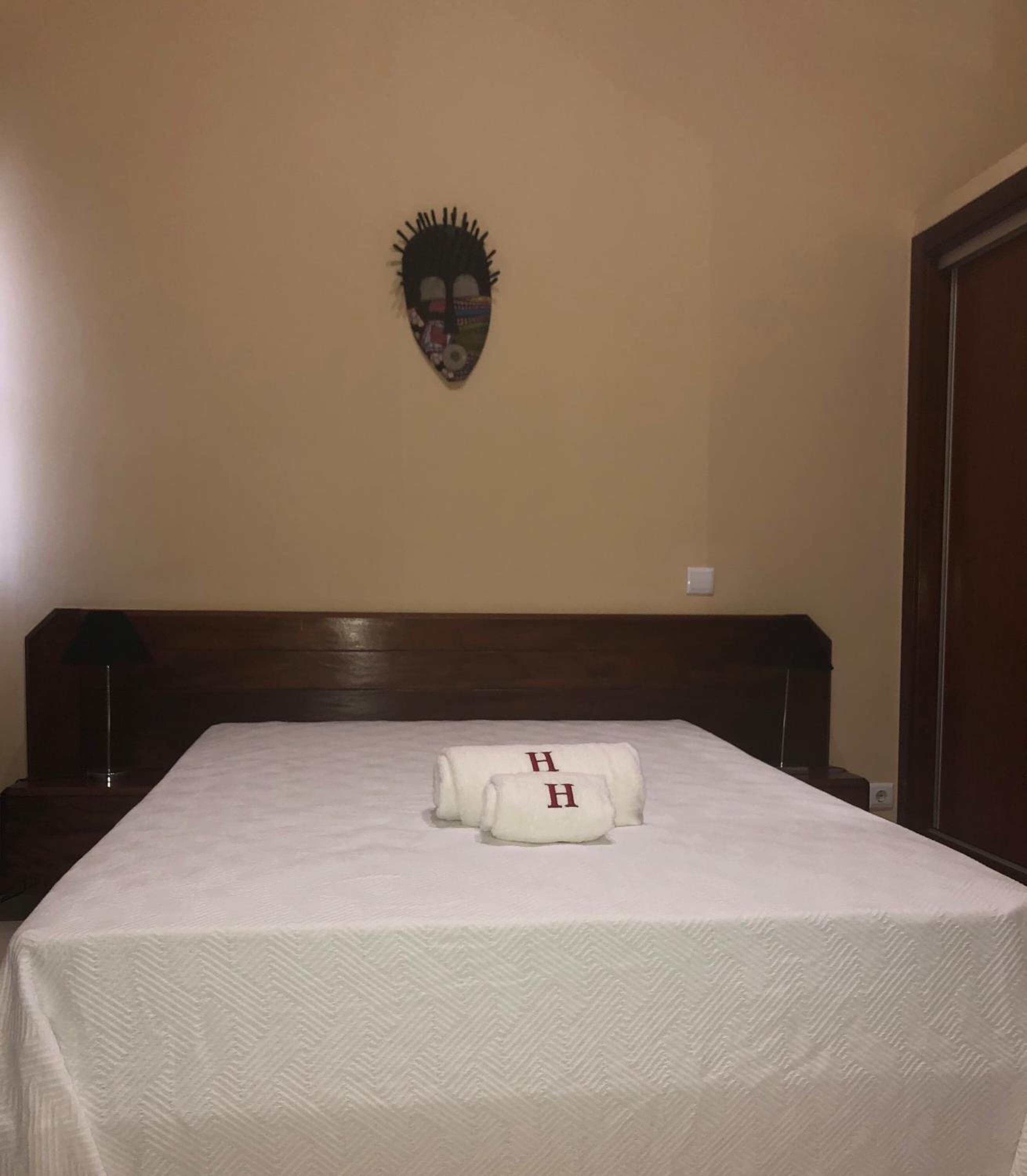 A hotel room with a bed, two towels with 'H' initials, a wooden headboard, and a wall decoration.