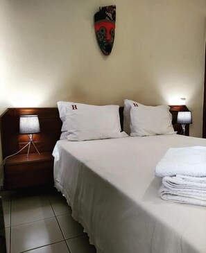 A hotel room with a bed, two pillows, a headboard, a nightstand with a lamp, and a wall-mounted decorative item. - Hotel Central (São Tomé)