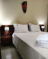 Habitación de hotel con cama, dos almohadas, cabecero, mesita de noche con lámpara y un adorno en la pared.