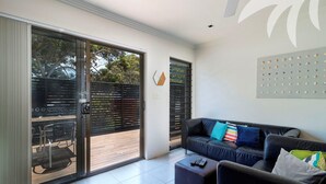 A modern living room with a black leather sofa, a wooden coffee table, and a sliding glass door leading to an outdoor area. - Aqua Apartment #1 - Pacific Palms Holidays (Blueys Beach)