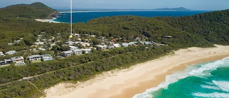 Luchtbeeld van een kustgebied met een strand, een dicht bos en een woongebied met huizen.