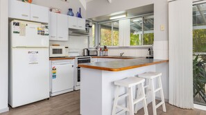 A modern kitchen with white cabinetry, a built-in oven, a microwave, and a coffee maker. There are two white bar stools and a window with a view of greenery outside. - Twin Peaks 1 at Boomerang Beach (Boomerang Beach)
