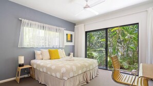 Une chambre à coucher comprenant un lit, une table de chevet, une chaise, une fenêtre avec des rideaux et une vue sur la verdure à l’extérieur.