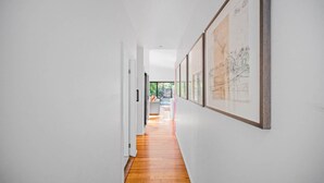 Un couloir aux murs blancs et au plancher en bois, qui mène à une pièce offrant une vue sur la verdure environnante.