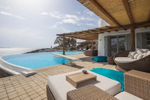 A poolside area with a wooden pergola, wicker furniture, and a view of the sea.