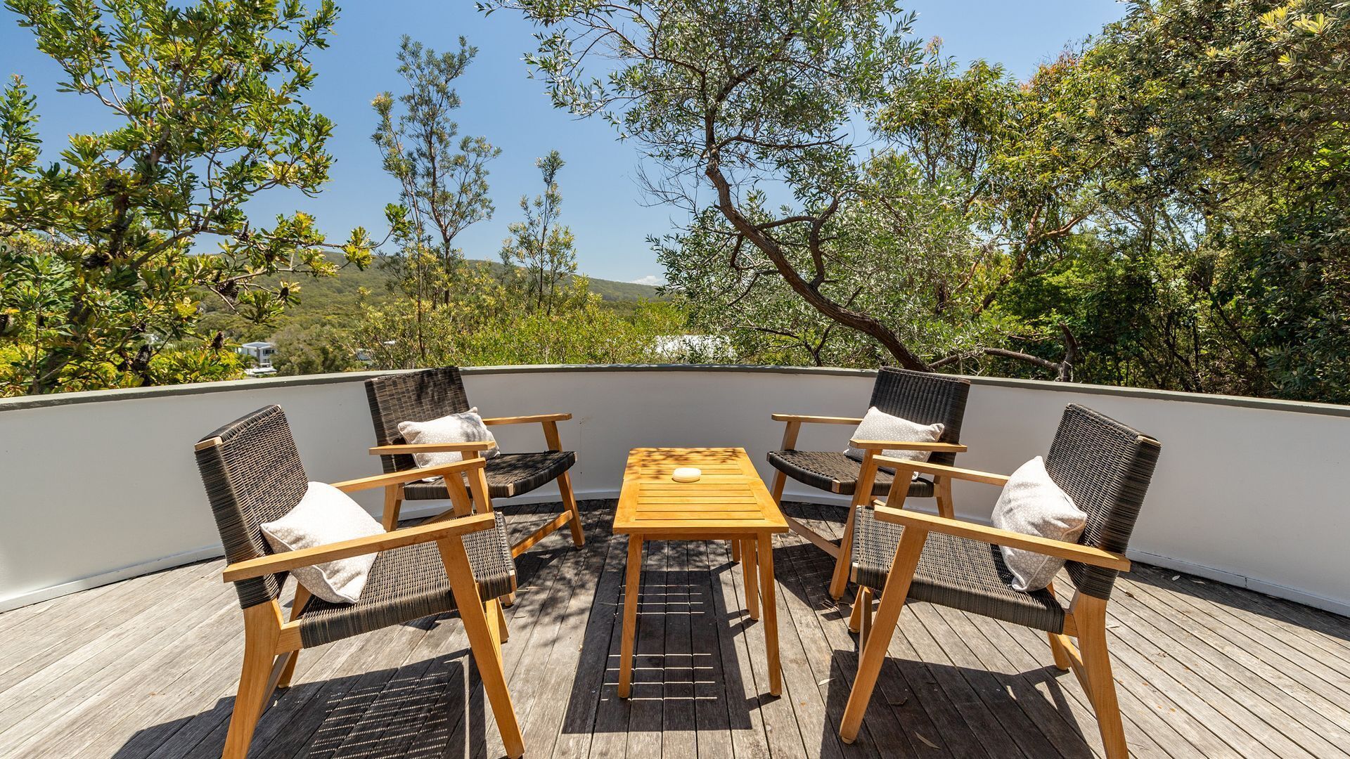 Una terraza con cuatro sillas de mimbre y una pequeña mesa de madera, rodeada por un muro blanco y con vistas a un paisaje exuberante.