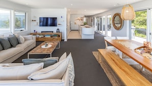A modern living room with a sofa, coffee table, and dining area. There is a TV mounted on the wall, a kitchen in the background, and large windows providing natural light. - Ningaloo - Boomerang Beach (Boomerang Beach)