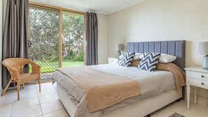 A bedroom with a large bed, a nightstand, a wicker chair, and a view of the outdoors through a glass door. - Oceanside 17  -  Breezes (Boomerang Beach)