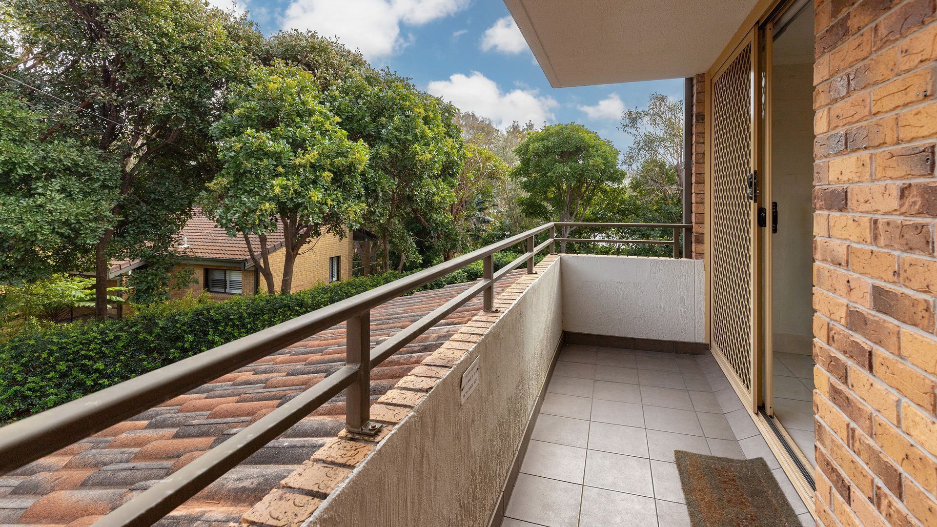 A balcony with a metal railing, a tiled floor, and a view of trees and a building in the background.