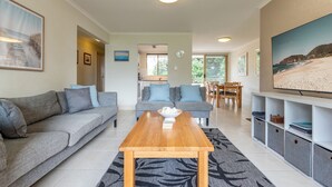A modern living room with a grey sofa, a wooden coffee table, and a dining area with a wooden table and chairs. - Oceanside 17  -  Breezes (Boomerang Beach)