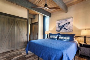 Une chambre avec un grand lit, des poutres apparentes en bois au plafond, un ventilateur de plafond, une grande fenêtre avec des stores, une œuvre d’art représentant une baleine accrochée au mur, et une table de chevet avec une lampe.