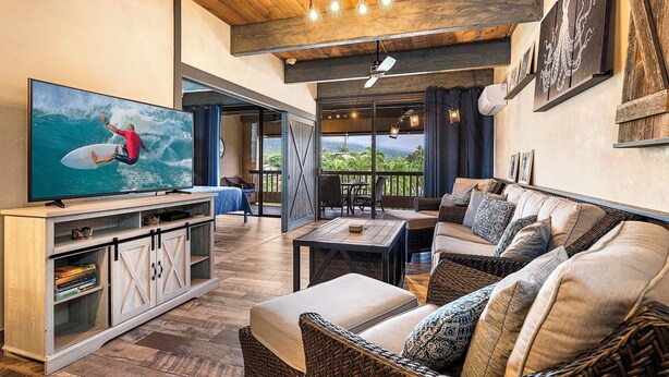 A spacious living area with a large flat-screen TV, wicker furniture, and a view of the outdoors. - Loft Style Completely Remodeled Condo (Kailua-Kona)