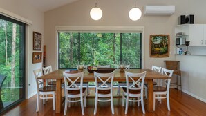 A dining area with a wooden table and white chairs, large windows with a forest view, and a kitchen area with cabinets and appliances. - A Yarraangunthi at Smiths Lake (Smiths Lake)