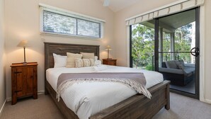 A bedroom with a large bed, wooden headboard, two bedside tables with lamps, a window with blinds, and a view of a balcony with outdoor furniture. - A Yarraangunthi at Smiths Lake (Smiths Lake)