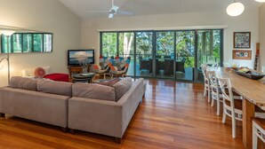 A modern living room with a sectional sofa, a dining area with a wooden table and chairs, a television, and a large sliding glass door leading to an outdoor patio. - A Yarraangunthi at Smiths Lake (Smiths Lake)