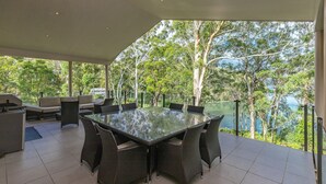A covered outdoor dining area with a glass table and wicker chairs, overlooking a lake and trees. - A Yarraangunthi at Smiths Lake (Smiths Lake)