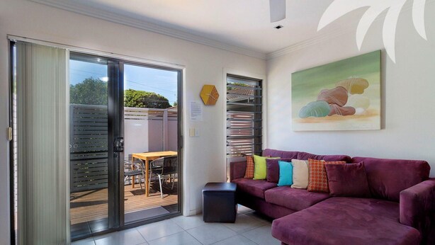 A living room with a purple sofa, a wooden table, and a glass door leading to an outdoor area. - Aqua Apartment #3 - Pacific Palms Holidays (Blueys Beach)