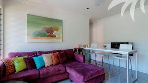 A modern living room with a purple sofa, colorful cushions, a dining area with a white table and metal chairs, and a painting on the wall. - Aqua Apartment #3 - Pacific Palms Holidays (Blueys Beach)