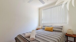 A bedroom with a bed, pillows, a bedside table, and a window with blinds. - Aqua Apartment #3 - Pacific Palms Holidays (Blueys Beach)