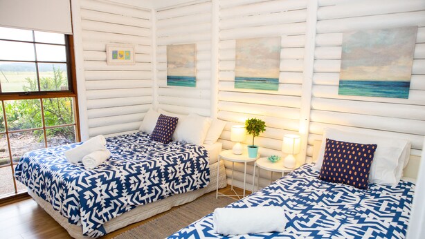 A room with two beds, each with a patterned bedspread, a small table with a lamp, and a view of the outdoors through a large window. - Large villa on private vineyard in prime and secluded location (Pokolbin)
