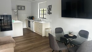 A modern kitchen with white cabinets, a dishwasher, and a window. There is a dining area with a glass table and four chairs. - Aqua Apartment #4 - Pacific Palms Holidays (Blueys Beach)