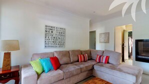 A living room with a beige sectional sofa, colorful pillows, a wooden side table, a wall-mounted TV, and a large sign listing various cities. - Aqua Apartment #4 - Pacific Palms Holidays (Blueys Beach)