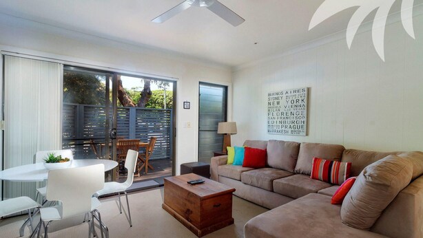 A modern living room with a sofa, coffee table, and chairs. There is a sliding glass door leading to an outdoor area with a table and chairs. A word cloud artwork is on the wall. - Aqua Apartment #4 - Pacific Palms Holidays (Blueys Beach)