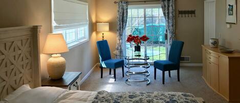 A hotel room with a bed, a desk with a lamp, two chairs, a ceiling fan, a window with curtains, and a view of the outdoors.