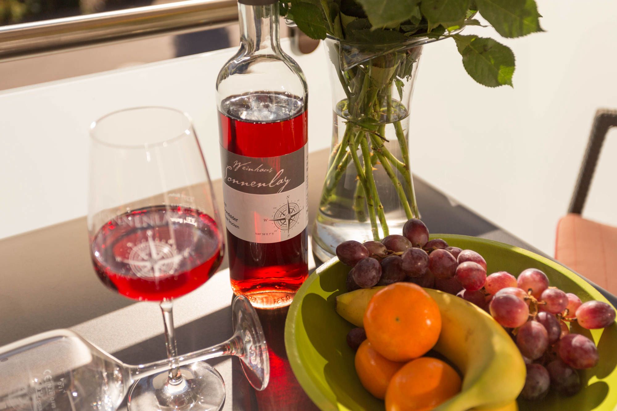 A table with a wine bottle, a glass of red wine, a vase with roses, and a bowl of fruit.