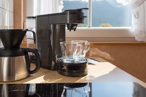 Une machine à café, une bouilloire et deux verres sont posés sur un plan de travail de cuisine, près d’une fenêtre offrant une vue sur les montagnes.
