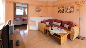 Un salon chaleureux avec un canapé, une table basse en bois et un téléviseur. On aperçoit un lit au fond et un tableau accroché au mur.