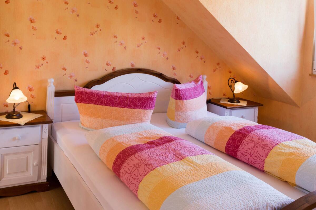 Een slaapkamer met een schuine kap, bloemenbehang, een houten hoofdbord, twee nachtkastjes met lampen, en een bed met een kleurrijke sprei.